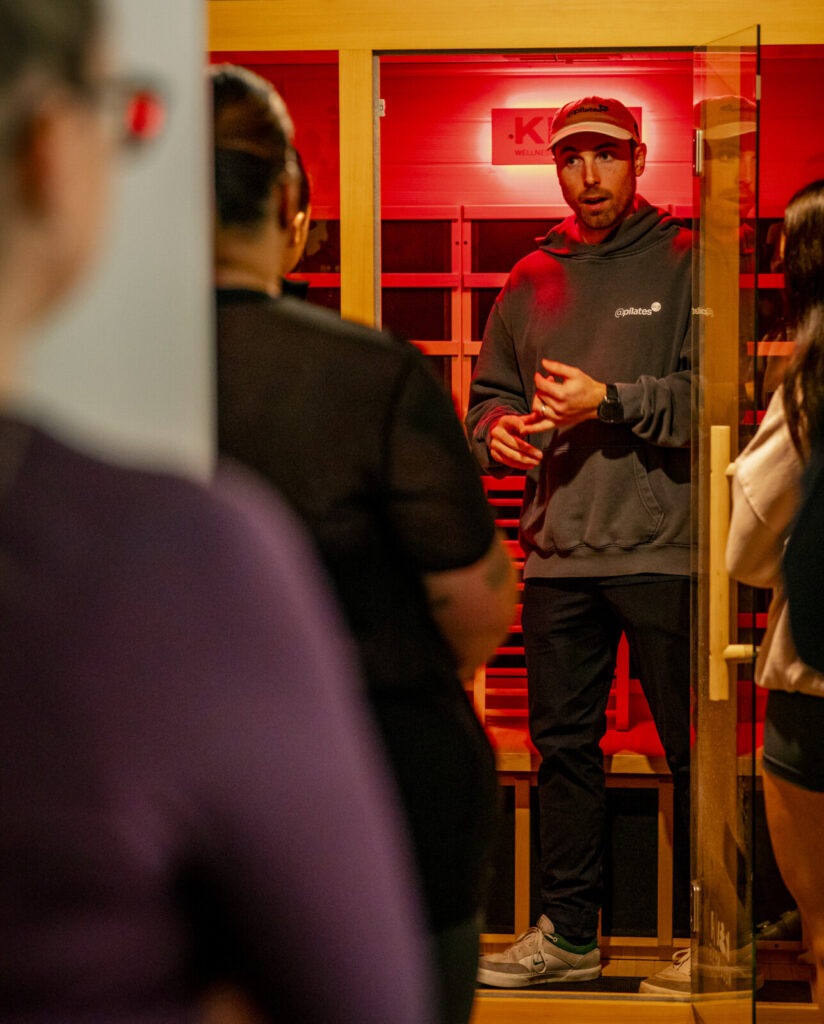 Man in red cap and black hoodie speaking to studio members in front of red neon signage at Pilates studio