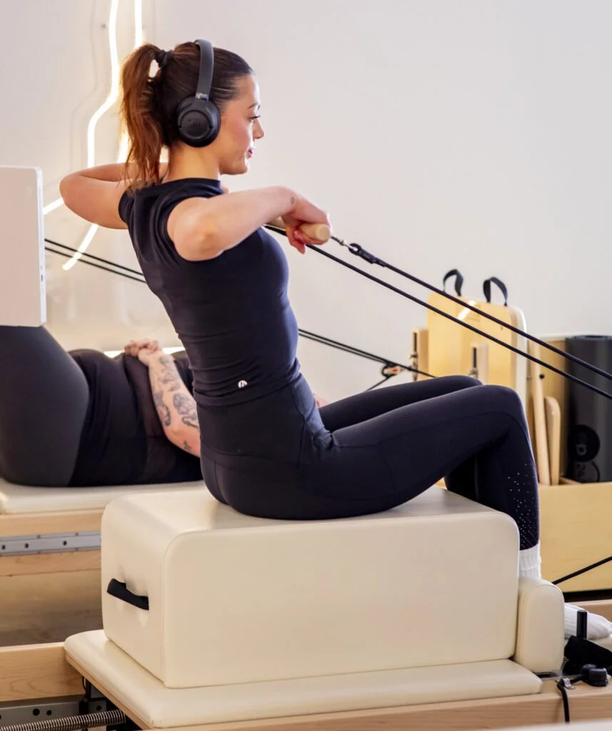 Woman on Pilates reformer wearing headphones, pulling resistance straps during a workout