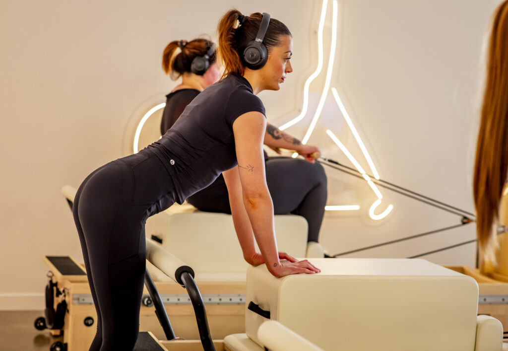 Woman in black workout gear wearing headphones performs a plank exercise on a Pilates reformer machine in a bright studio