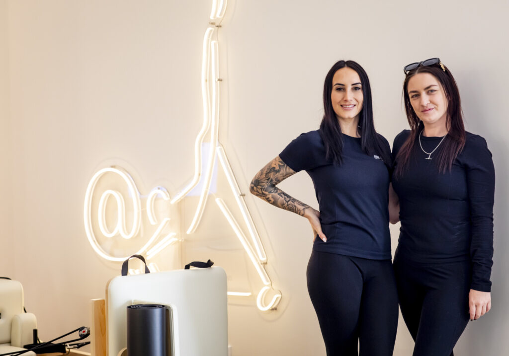 Two women in black athletic wear stand smiling next to a neon light display shaped like a guitar pick in the studio