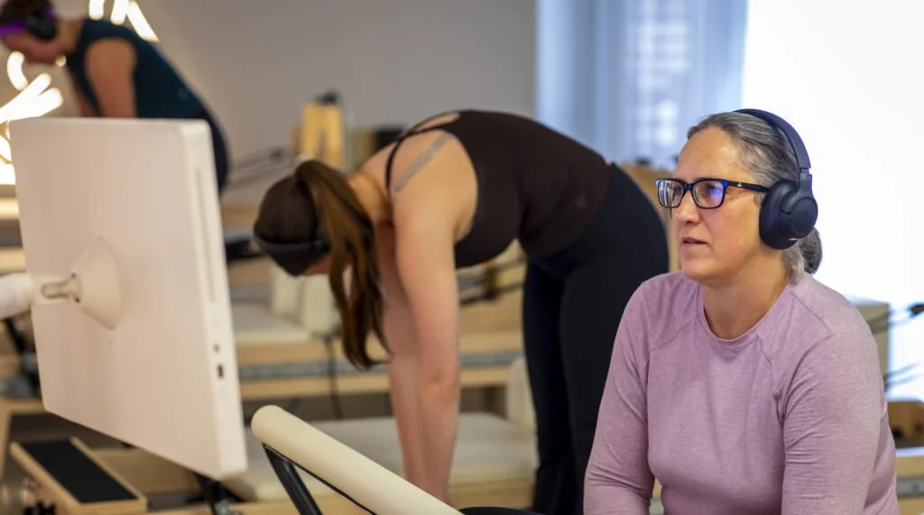 Woman wearing headphones and purple shirt seated at studio desk, with another person doing a pilates exercise in background